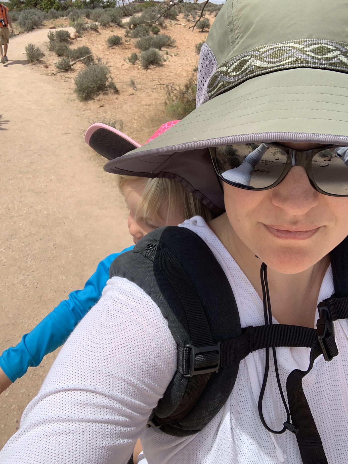 Laurel hiking in 2019, wearing a wide sun hat and sunglasses, carrying her young daughter in a back carrier on a desert trail.